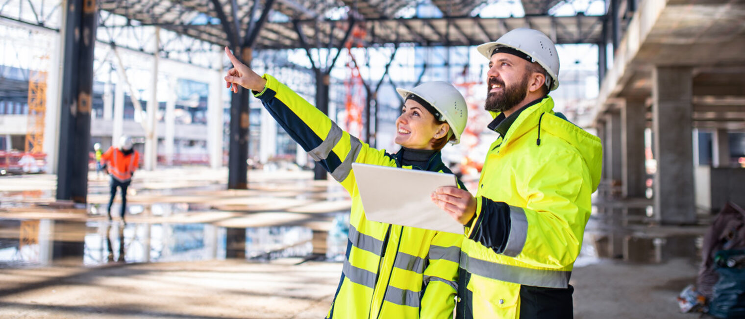 zwei Bauarbeitende lesen eine Planskizze und zeigen die geplanten Bauprojekte auf.