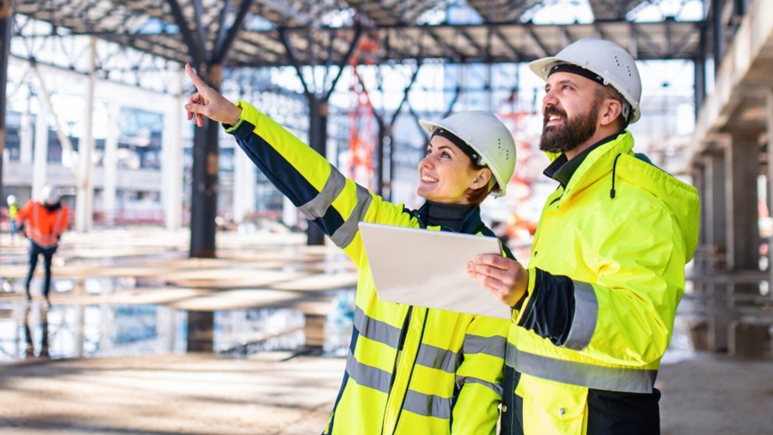 zwei Bauarbeitende lesen eine Planskizze und zeigen die geplanten Bauprojekte auf.