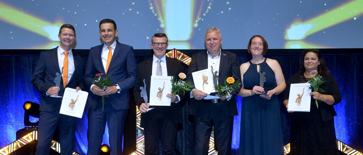 Drei Betriebs-Finalisten des Mittelstandspreises stehen auf der Bühne und halten ihren Pokal in der Nacht. 