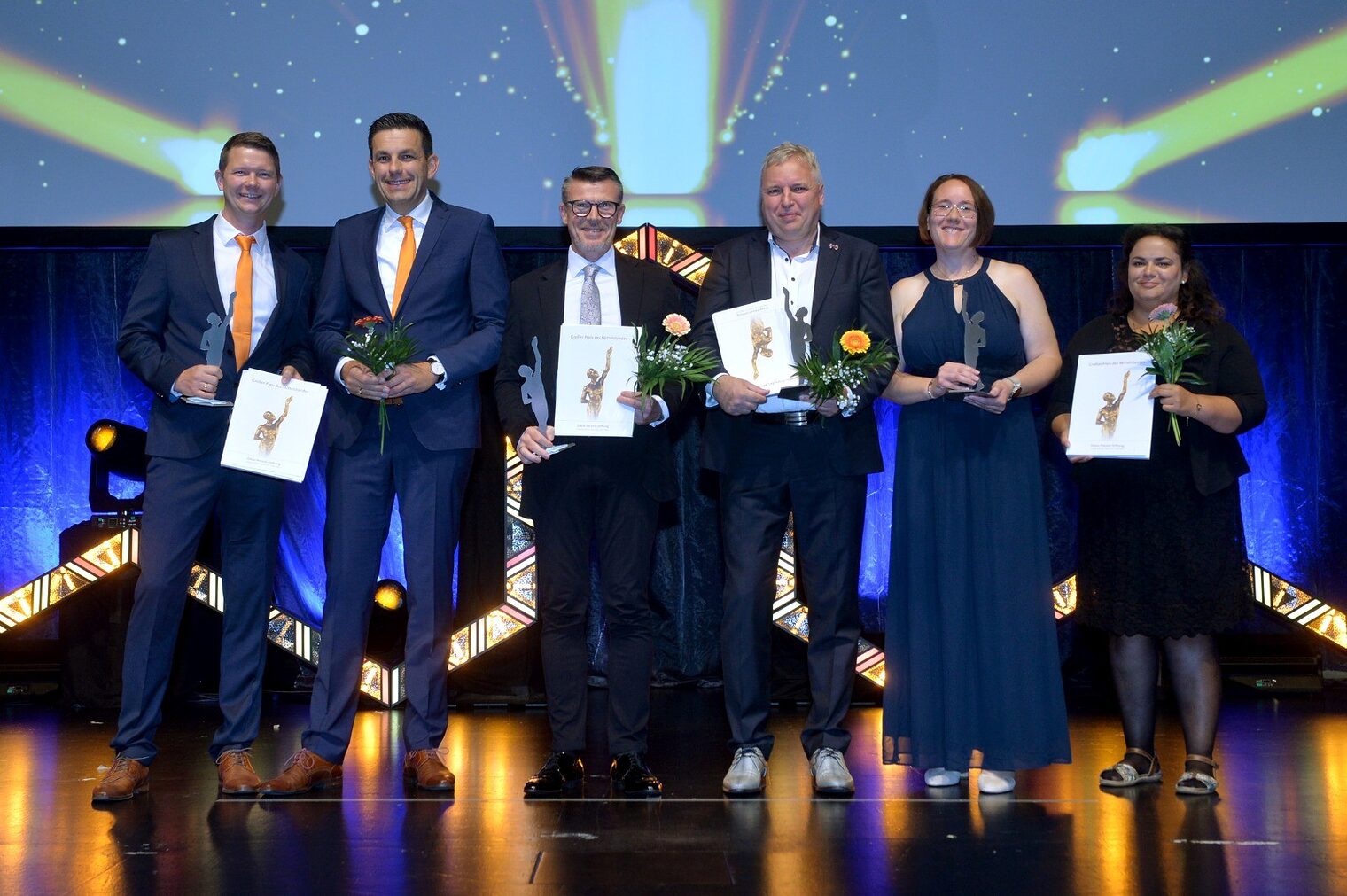 Drei Betriebs-Finalisten des Mittelstandspreises stehen auf der Bühne und halten ihren Pokal in der Nacht. 